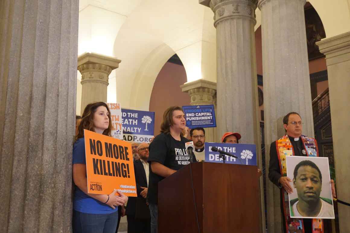 Anti-death penalty activists including clergy stand behind a lectern, beside some columns inside the South Carolina State House lobby.