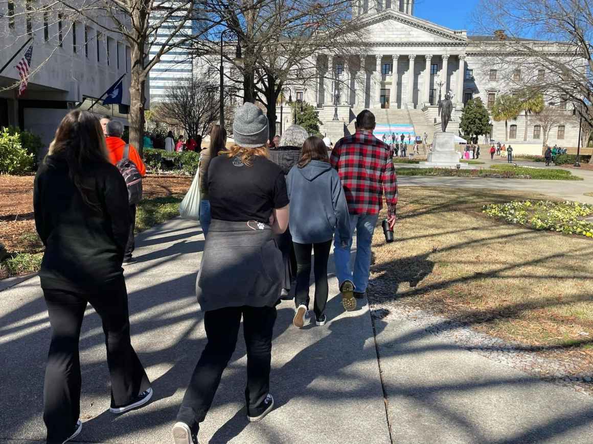 Eric and his family, backs toward the camera, walking toward the South Carolina State House. In the distance, a giant trans pride flag can be seen draped on the State House steps.