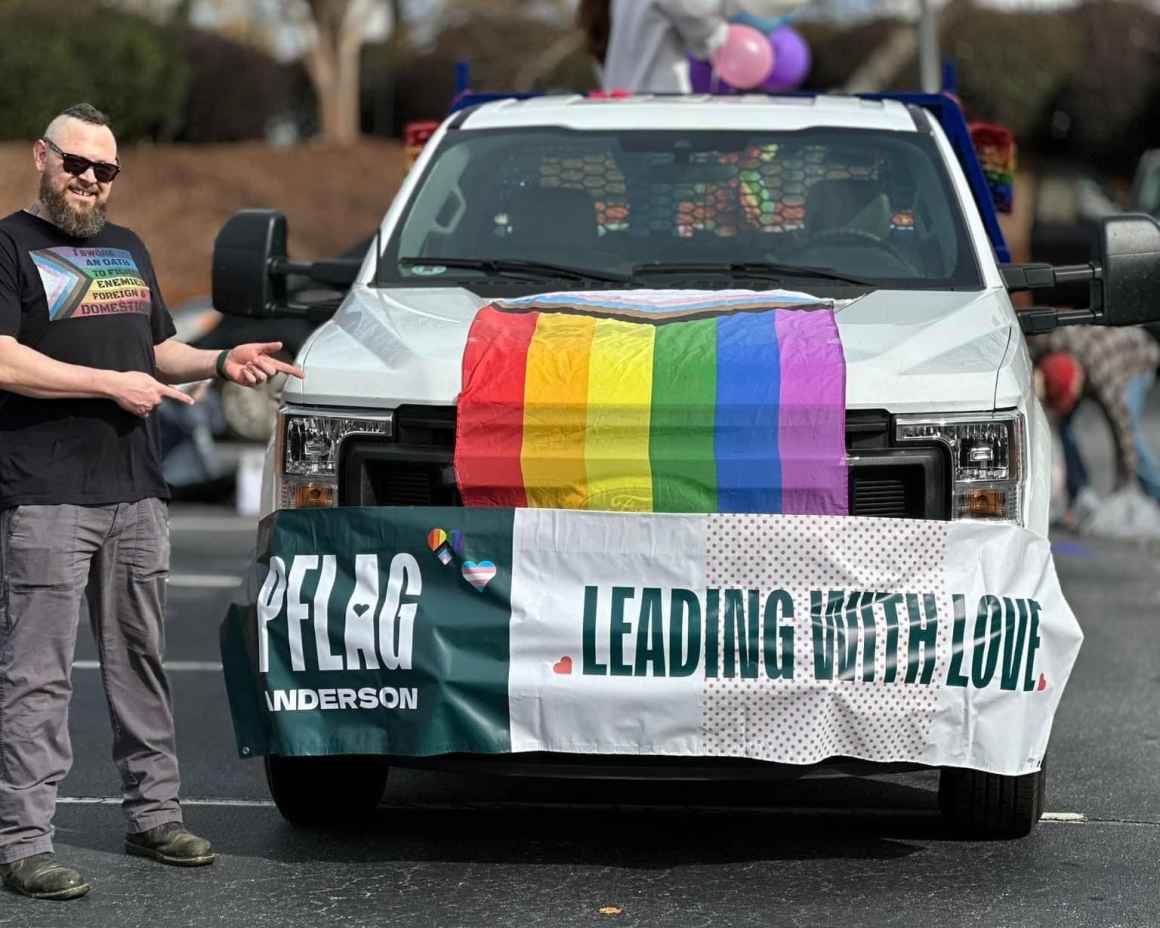 Eric standing beside a pickup truck preparing for a Pride parade. The truck has a rainbow flag and a PFLAG Anderson banner on the front with the slogan "Leading with love."