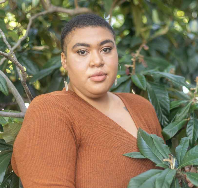 Portrait photo of Demetria standing surrounded by the branches of a tree with waxy broad leaves. Demetria is wearing a brown top and a neutral expression.