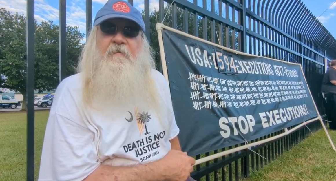 A man with a long white beard sits beside a black banner that reads, "USA: 1594 Executions 1977 - Present. Stop Executions!"