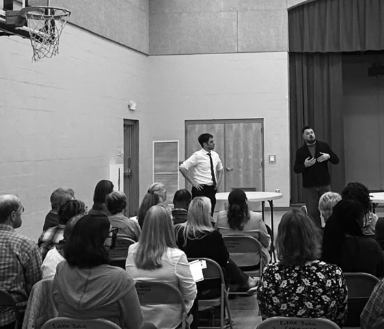 A crowd in a gymnasium listening to two speakers